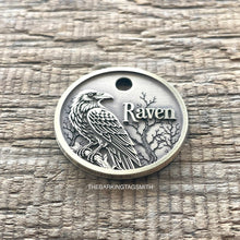 Load image into Gallery viewer, A round silver metal pet ID tag featuring an engraved raven, bare tree branches, and the word 'Raven' on a weathered wood background.