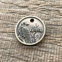Load image into Gallery viewer, A round silver-toned metal pet tag engraved with a detailed raven, bare tree branches, and the word 'Raven,' photographed on a textured wood surface. A small watermark reading THEBARKINGTAGSMITH appears near the bottom.