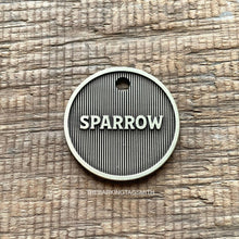 Load image into Gallery viewer, Macro product photograph of a circular, light champagne brushed brass pet tag with the name "SPARROW" in raised 3D lettering. The design features a deep fiber-laser engraved background of vertical pinstripe machined lines with a dark oxidized charcoal finish. The tag is centered on a weathered, brown horizontal-grain wooden surface with visible cracks and natural lighting.