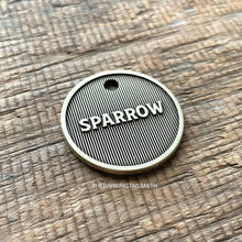 Load image into Gallery viewer, Macro product photograph of a circular, light champagne brushed brass pet tag with the name "SPARROW" in raised 3D lettering. The design features a deep fiber-laser engraved background of vertical pinstripe machined lines with a dark oxidized charcoal finish. The tag is centered on a weathered, brown horizontal-grain wooden surface with visible cracks and natural lighting.