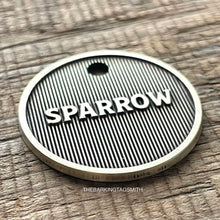 Load image into Gallery viewer, Macro product photograph of a circular, light champagne brushed brass pet tag with the name "SPARROW" in raised 3D lettering. The design features a deep fiber-laser engraved background of vertical pinstripe machined lines with a dark oxidized charcoal finish. The tag is centered on a weathered, brown horizontal-grain wooden surface with visible cracks and natural lighting.