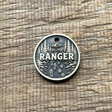 Load image into Gallery viewer, Personalised forest trail dog ID tag with engraved mountain landscape, pine trees, hiking boot prints and paw prints in brushed silver metal on rustic wood background