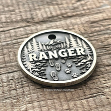 Load image into Gallery viewer, Personalised forest trail dog ID tag with engraved mountain landscape, pine trees, hiking boot prints and paw prints in brushed silver metal on rustic wood background
