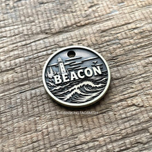 Load image into Gallery viewer, Personalised lighthouse dog ID tag with engraved ocean waves, coastal lighthouse and shoreline scene in brushed silver metal on rustic wood background